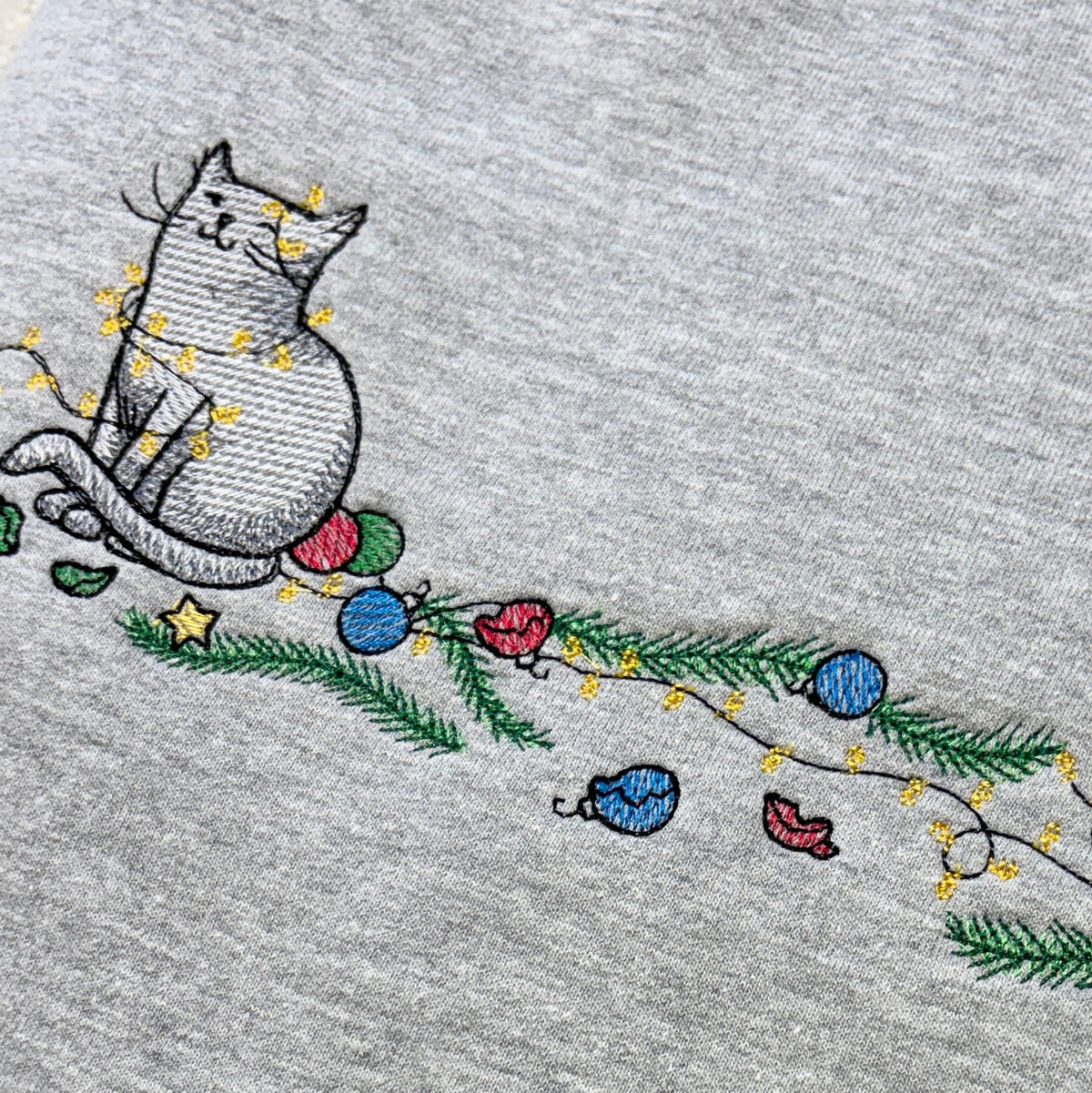 Folded grey sweatshirt with Cat embroidery with christmas tree baubles and lights strewn around it