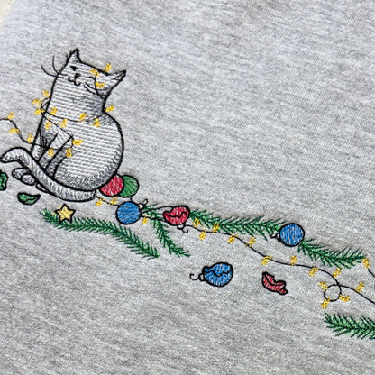 Folded grey sweatshirt with Cat embroidery with christmas tree baubles and lights strewn around it