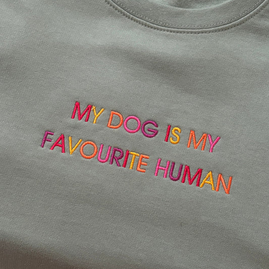 A close-up of a sage green crewneck t-shirt featuring colorful embroidered text that reads 'MY DOG IS MY FAVOURITE HUMAN' in pink, orange, and yellow.