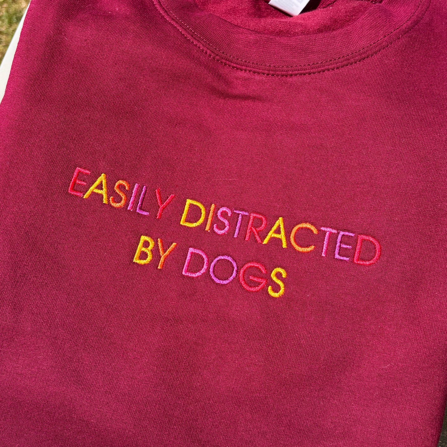 A folded maroon crewneck sweatshirt with the phrase 'EASILY DISTRACTED BY DOGS' embroidered across the chest in a rainbow of colors.