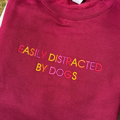 A folded maroon crewneck sweatshirt with the phrase 'EASILY DISTRACTED BY DOGS' embroidered across the chest in a rainbow of colors.