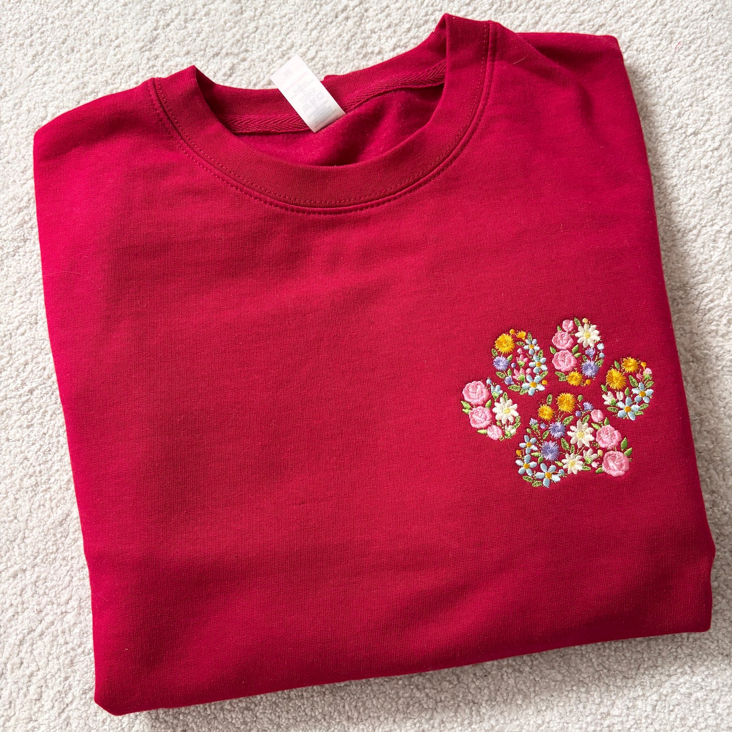 A folded red crewneck sweatshirt featuring a colorful embroidered floral paw print on the left chest, laid flat on a textured beige carpet.