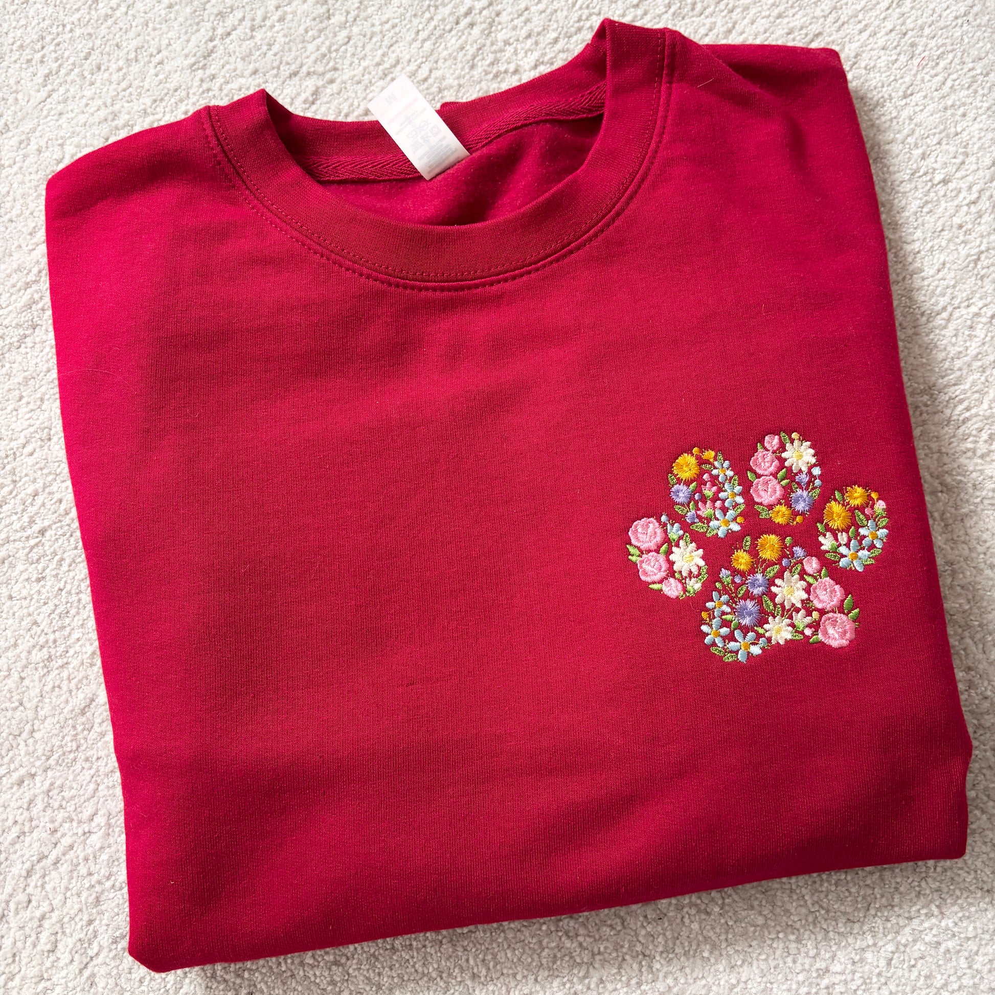 A folded red crewneck sweatshirt featuring a colorful embroidered floral paw print on the left chest, laid flat on a textured beige carpet.