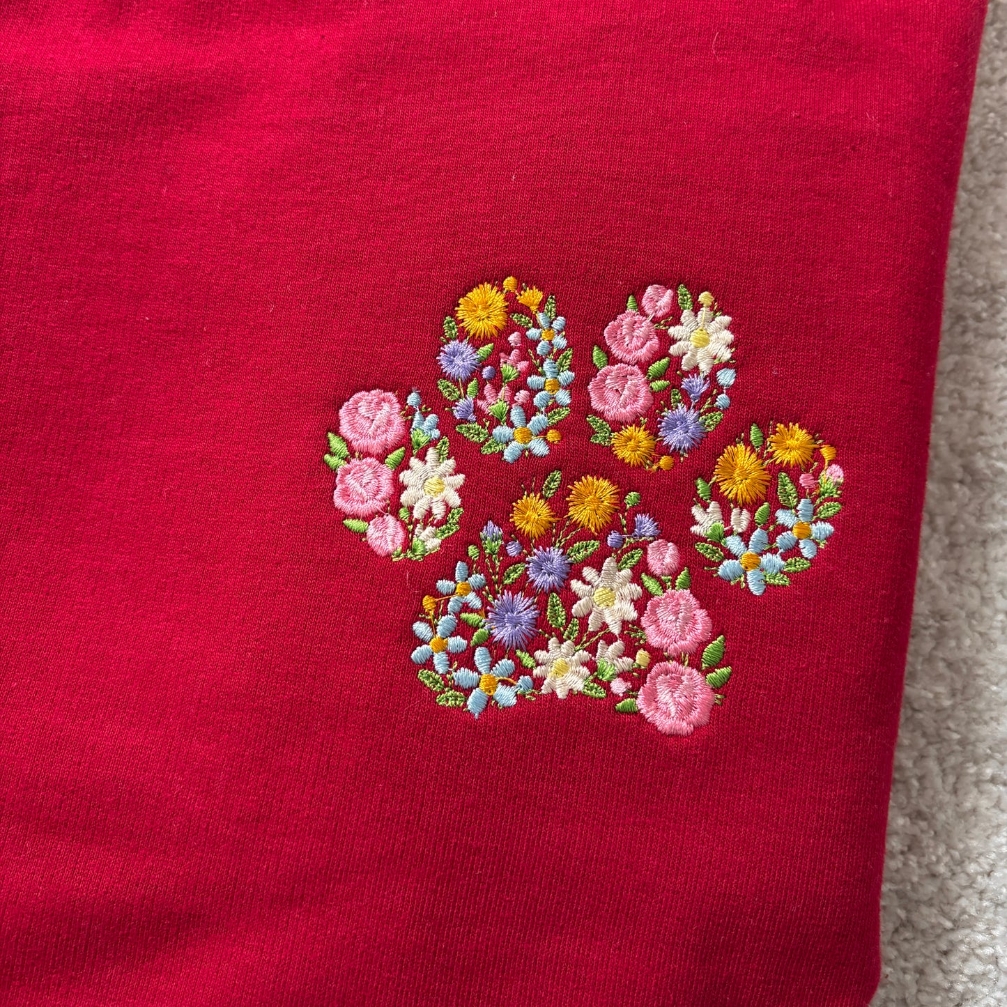 A close-up of a red knit garment featuring an embroidered paw print made from colorful flowers in pink, yellow, blue, and white, with green leaves.