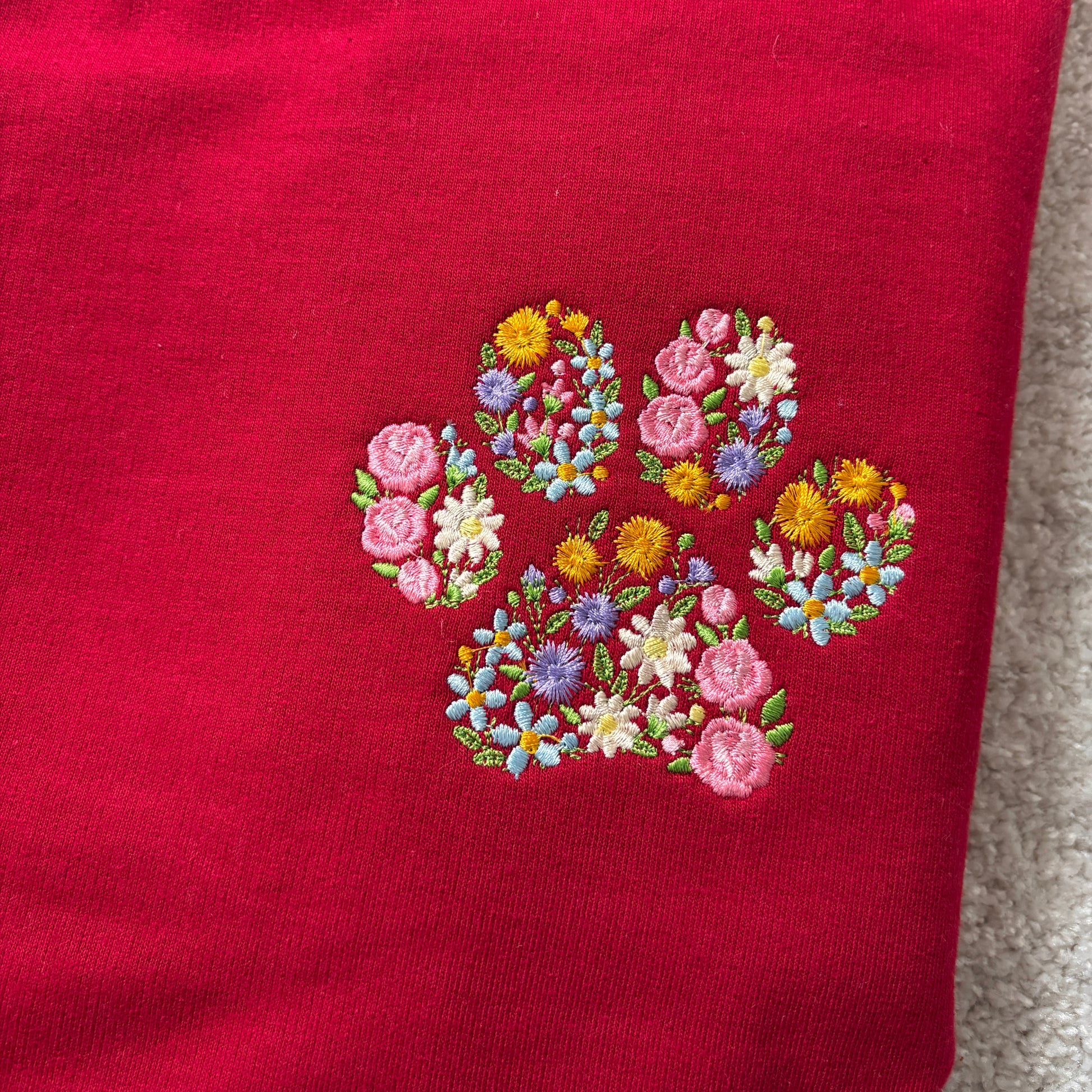 A close-up of a red knit garment featuring an embroidered paw print made from colorful flowers in pink, yellow, blue, and white, with green leaves.