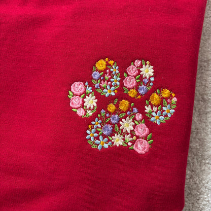A close-up of a red knit garment featuring an embroidered paw print made from colorful flowers in pink, yellow, blue, and white, with green leaves.