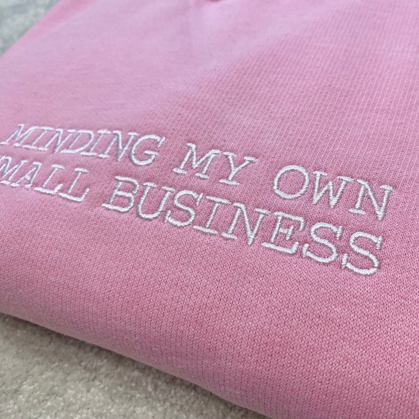 Pink fabric with embroidered text 'Minding My Own Small Business' on a sweatshirt
