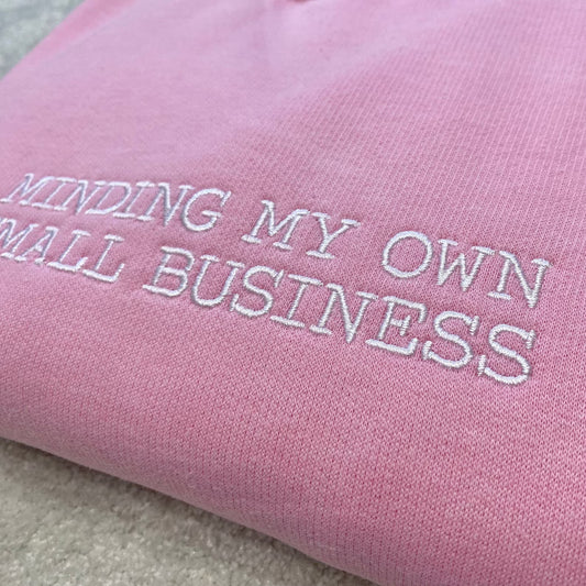 Pink fabric with embroidered text 'Minding My Own Small Business' on a sweatshirt