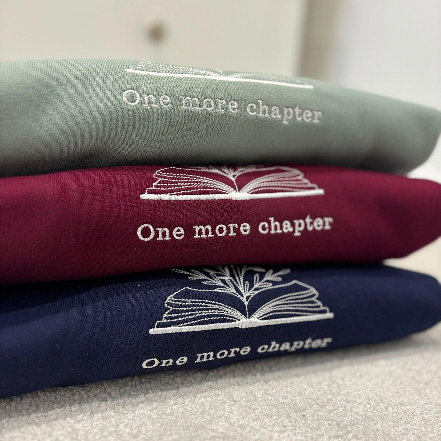 Stack of three sweatshirts with 'One more chapter' text and book illustration embroidered.