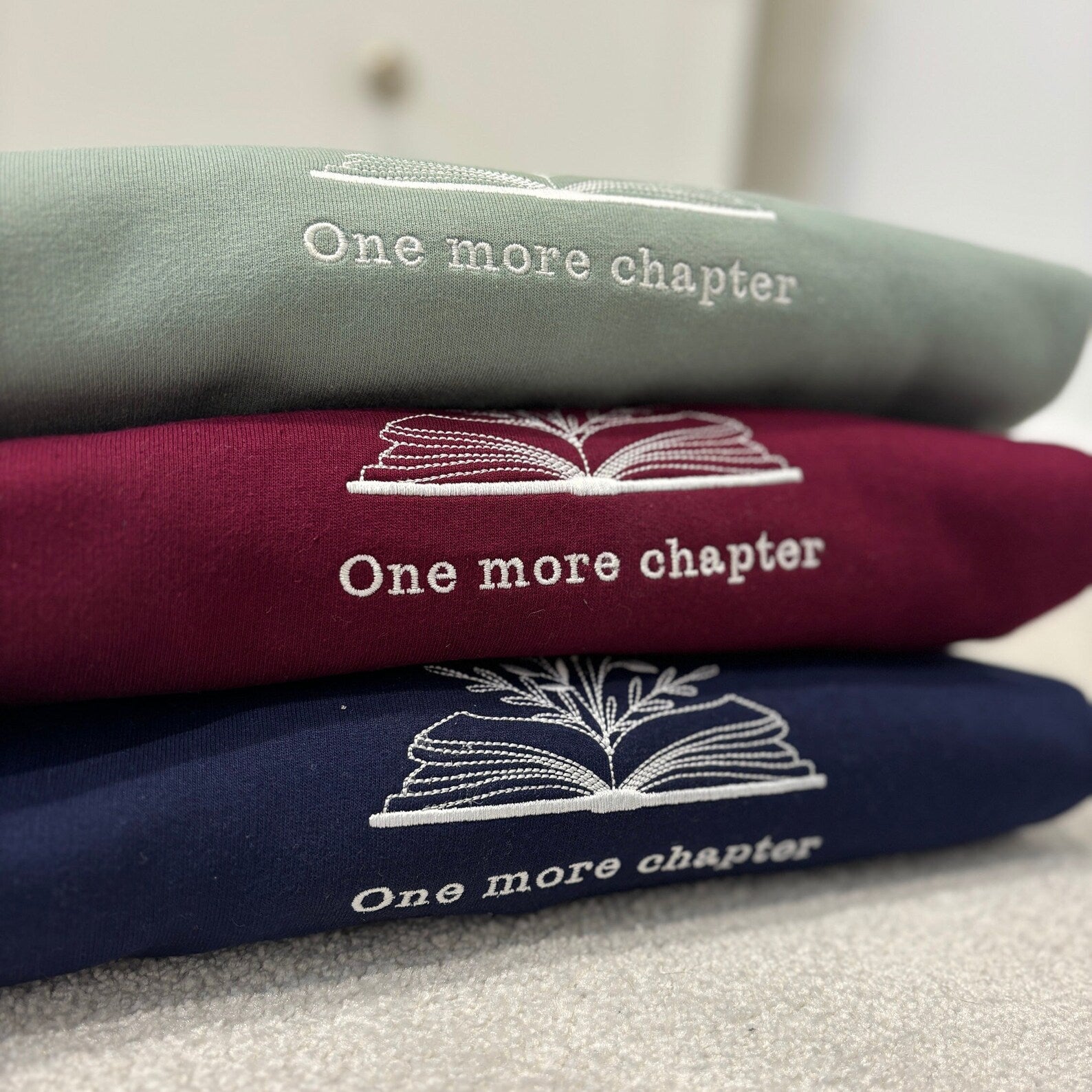 Stack of three sweatshirts with 'One more chapter' text and book illustration embroidered.