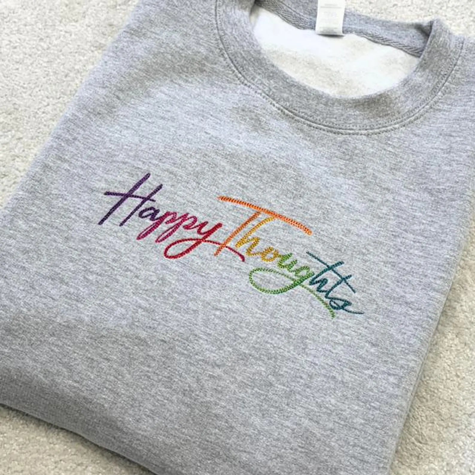 Happy Thoughts embroidered in rainbow colourful colours on a grey folded sweatshirt