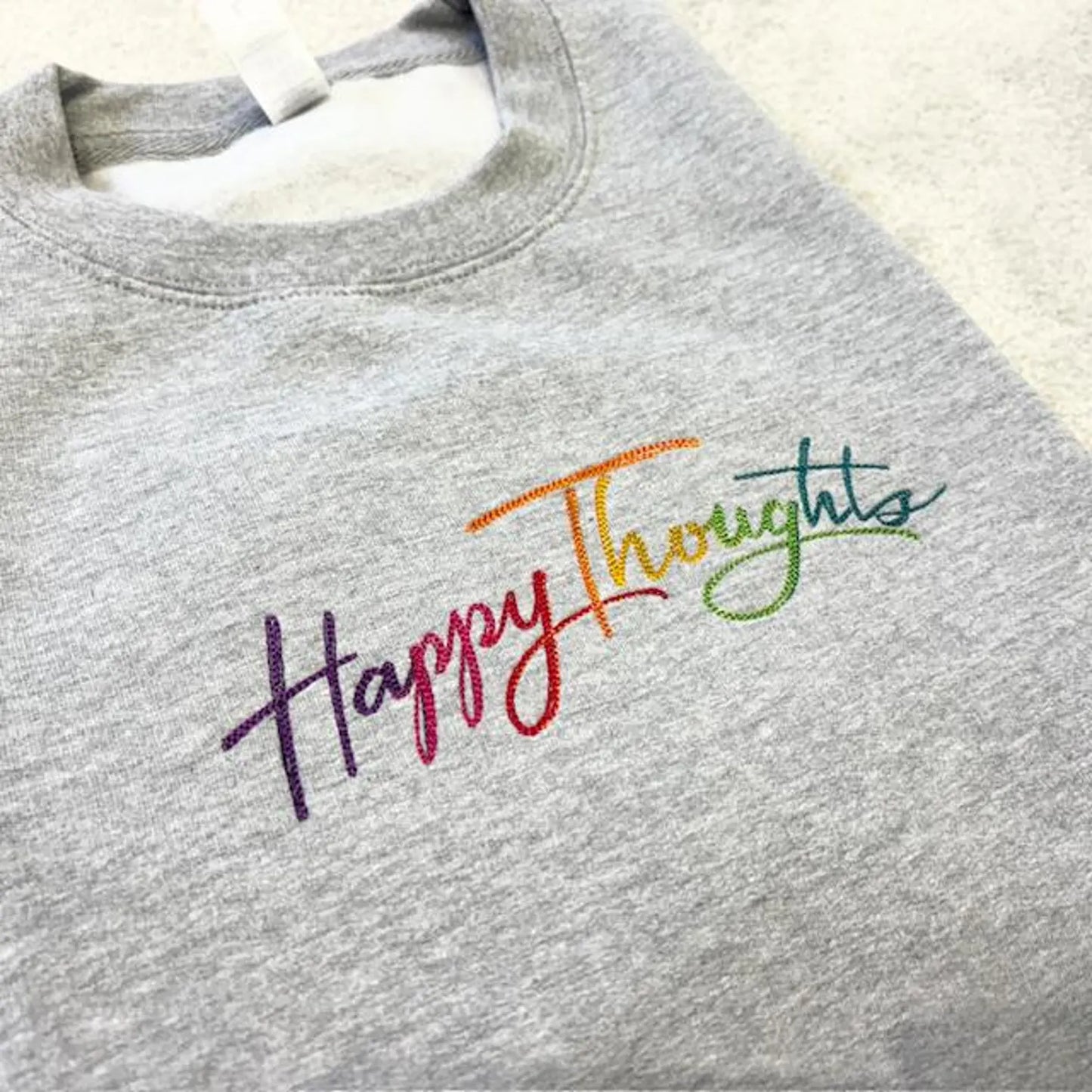 Happy Thoughts embroidered in rainbow colourful colours on a grey folded sweatshirt