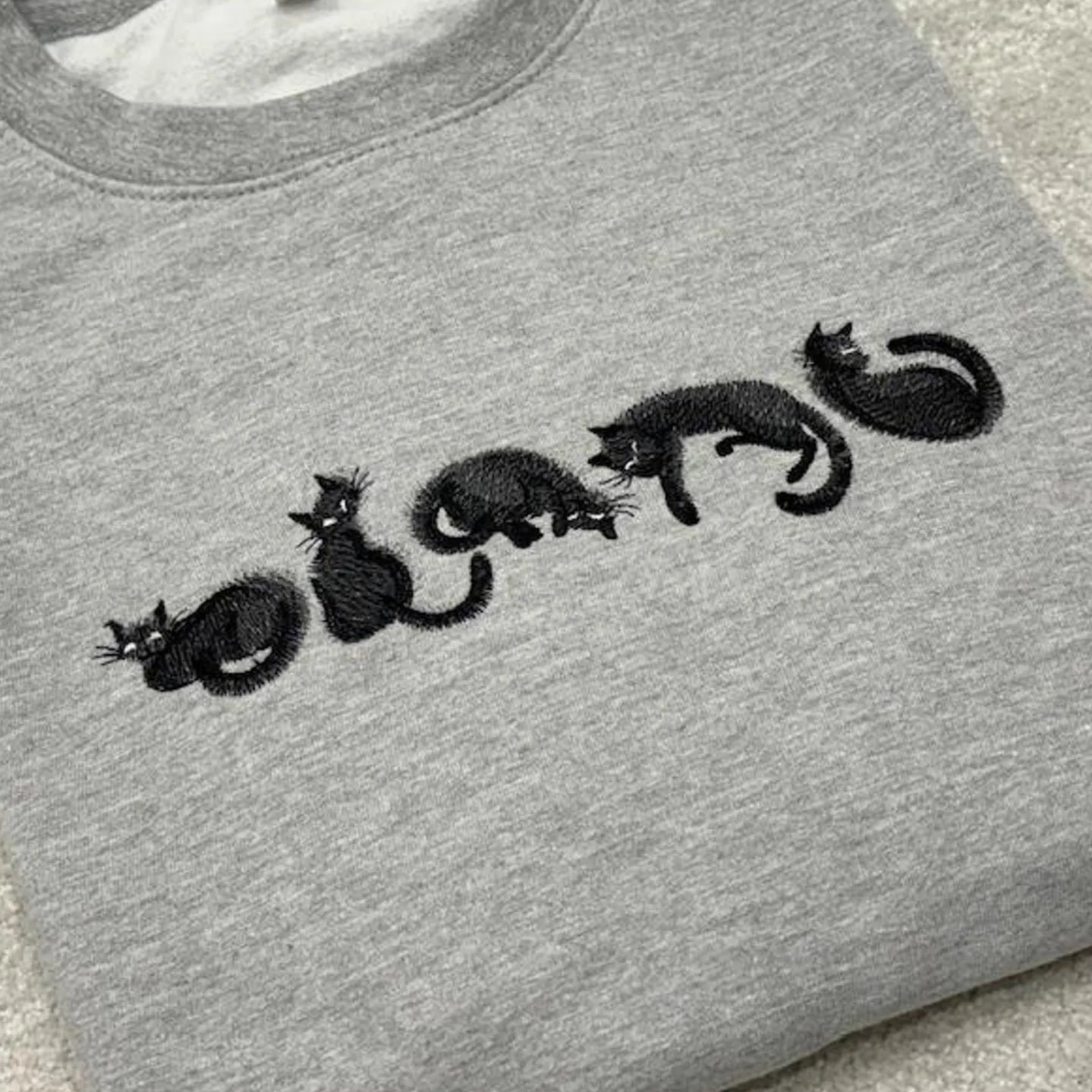 Grey sweatshirt with five black cats laying in a row, embroidered in a fuzzy stitch style.