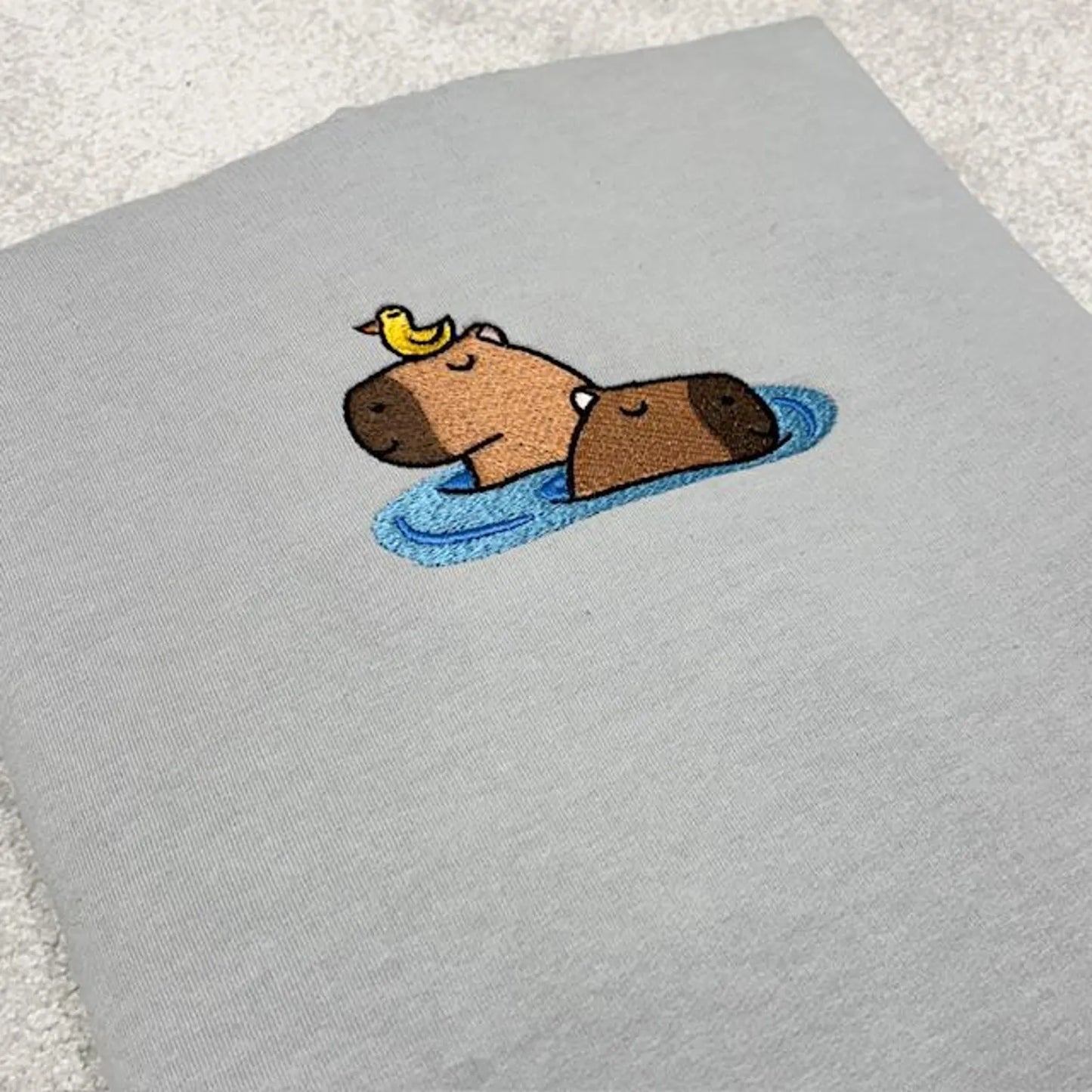 Embroidered design of a capybara and a bird on a grey fabric background