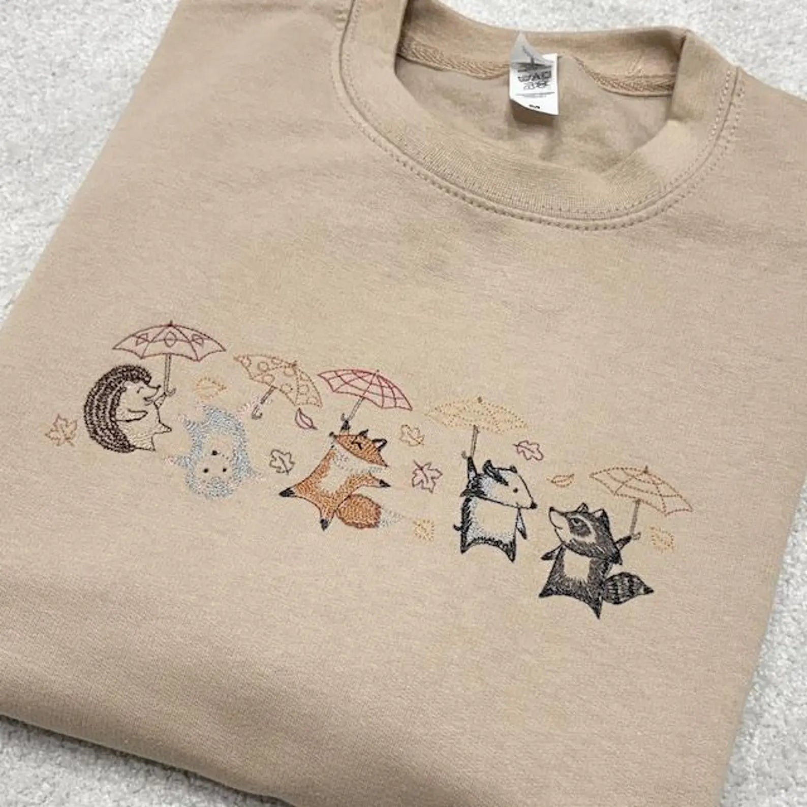 Woodland Animals fighting the blustering wind with umbrellas embroidered across the front of a sand coloured sweatshirt.