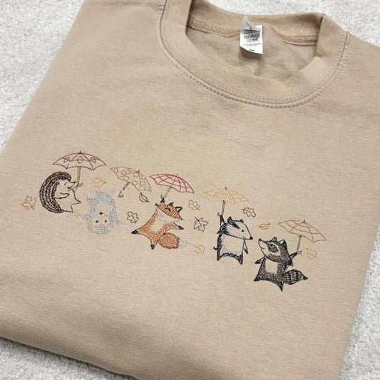 Woodland Animals fighting the blustering wind with umbrellas embroidered across the front of a sand coloured sweatshirt.