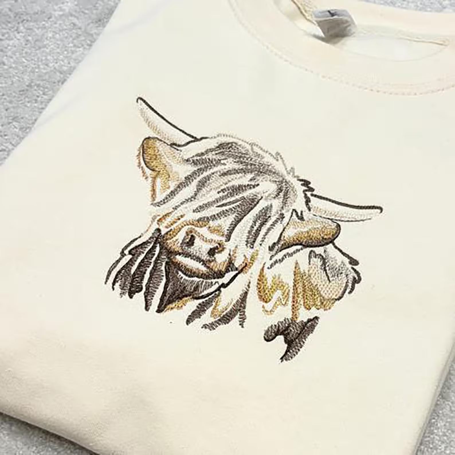 Embroidered design of a highland cow on a cream sweatshirt.